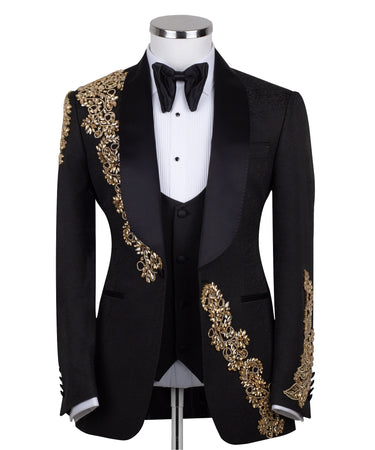 Front view of a black jacquard three-piece tuxedo with intricate gold hand-embellishments on the shawl lapel, shoulder, and cuffs, displayed on a mannequin. From BTS Bespoke, handcrafted in Istanbul.
