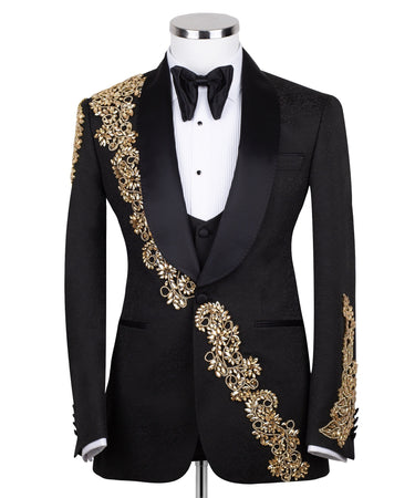Front view of a black jacquard three-piece tuxedo with intricate gold hand-embellishments on the shawl lapel, shoulder, and cuffs, displayed on a mannequin. From BTS Bespoke, handcrafted in Istanbul.