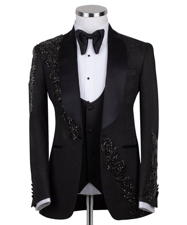 Black slim fit hand-embellished tuxedo on a mannequin, featuring a satin peak lapel, matching vest, and intricate black beadwork detailing on the shoulder, lapel, and sleeves. The tuxedo fabric has a subtle jacquard texture, showcasing master tailoring from Istanbul.