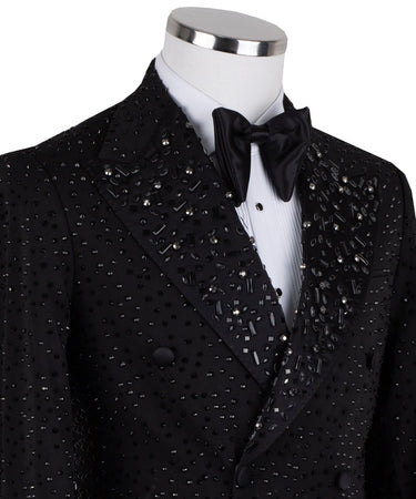 Close-up of a black double-breasted tuxedo jacket on a mannequin, showcasing intricate hand-embellishments with beads and crystals on the lapels and body. Features a peak lapel and black buttons, paired with a white shirt and black bow tie. Front view showing the full jacket and details.