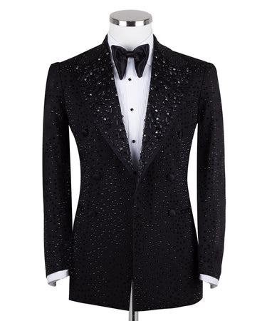 Close-up of a black double-breasted tuxedo jacket on a mannequin, showcasing intricate hand-embellishments with beads and crystals on the lapels and body. Features a peak lapel and black buttons, paired with a white shirt and black bow tie. Front view showing the full jacket and details.