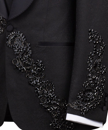 Black slim fit hand-embellished tuxedo on a mannequin, featuring a satin peak lapel, matching vest, and intricate black beadwork detailing on the shoulder, lapel, and sleeves. The tuxedo fabric has a subtle jacquard texture, showcasing master tailoring from Istanbul.