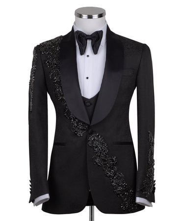 Black slim fit hand-embellished tuxedo on a mannequin, featuring a satin peak lapel, matching vest, and intricate black beadwork detailing on the shoulder, lapel, and sleeves. The tuxedo fabric has a subtle jacquard texture, showcasing master tailoring from Istanbul.