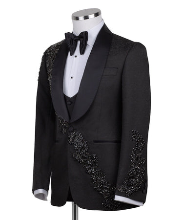 Black slim fit hand-embellished tuxedo on a mannequin, featuring a satin peak lapel, matching vest, and intricate black beadwork detailing on the shoulder, lapel, and sleeves. The tuxedo fabric has a subtle jacquard texture, showcasing master tailoring from Istanbul.