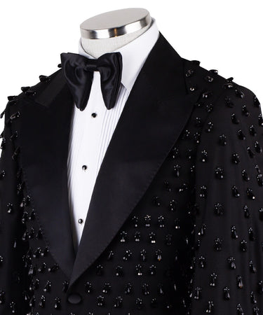 Front view of a black hand-embellished luxury tuxedo jacket with satin peak lapels and matching trousers on a mannequin. Side view close-up of the jacket's intricate black bead embellishments. Close-up of the tuxedo trousers with a satin side stripe and high-waisted detail.